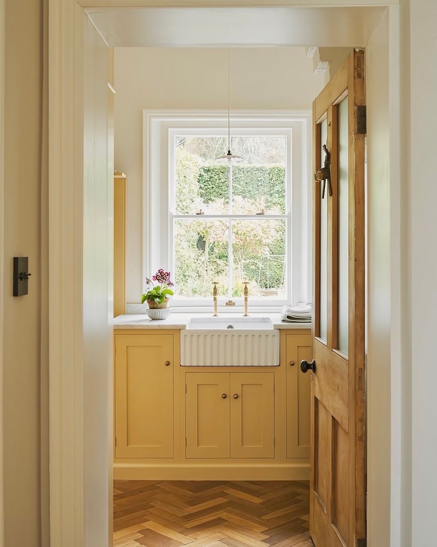 A pastel yellow kitchen, centered by a white belfast sink with golden taps, set below a sash window